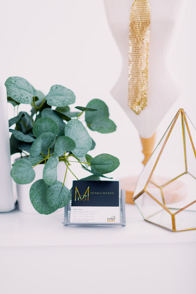 Realtor Business Cards, a Plant, a Miniature Fasion Mannequin with a Sparkly Tie, and an Empty Geometric Glass Terrarium are all Positioned on a White Desk