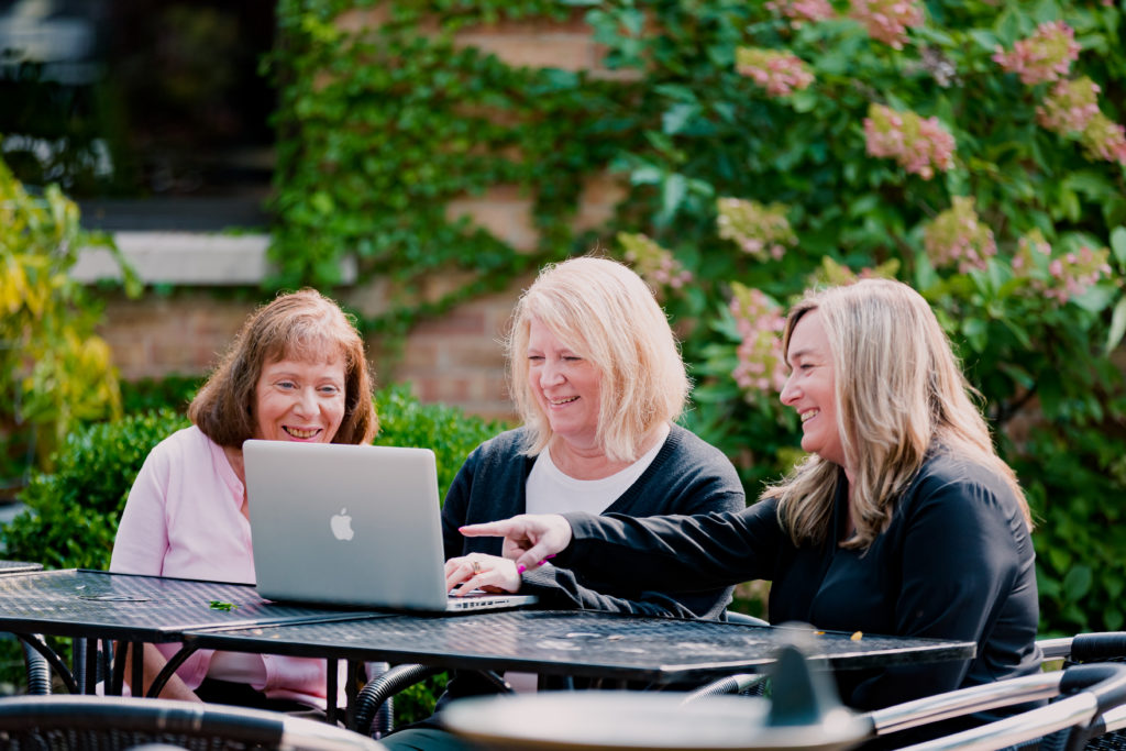 Three Coworkers Smiling and Pointing at a Laptop on Patio Furniture
