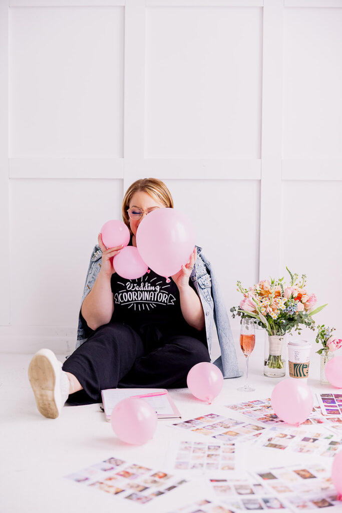 Vibrant and fun brand photo of a woman sitting holding pink balloons in front of her face, surrounded by flowers and mood boards, taken by This Way to Fabulous