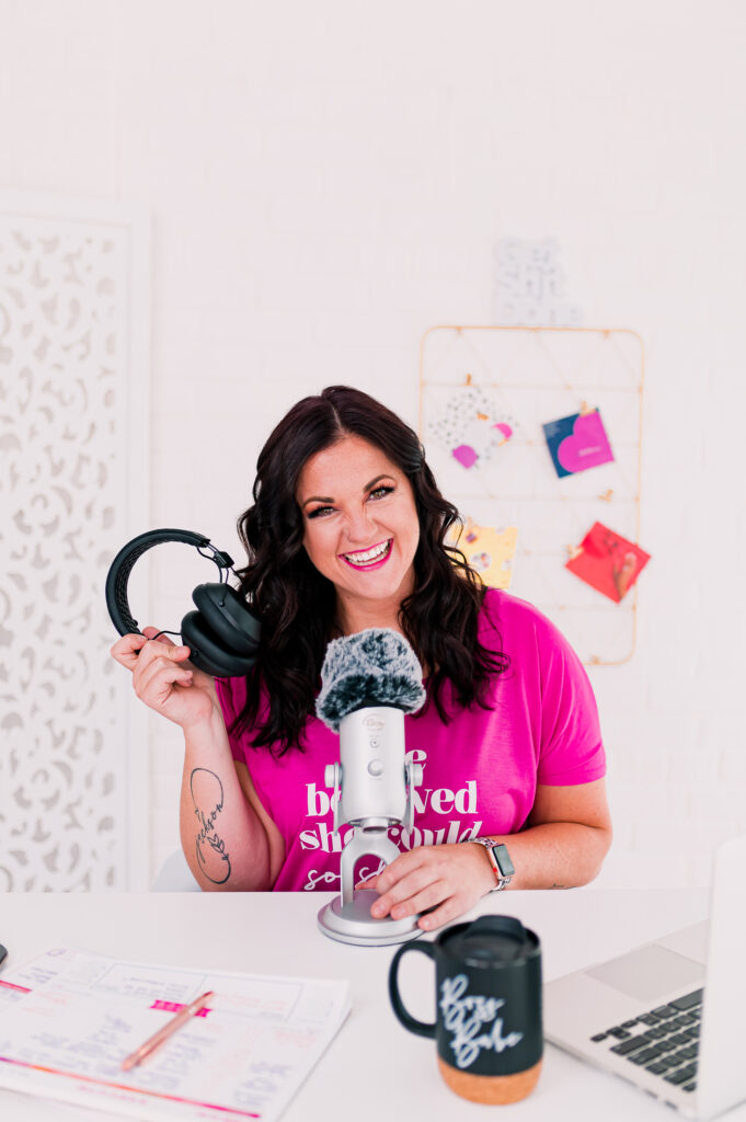 Headshot-style branding photo of Jessi from This Way to Fabulous holding headphones and a microphone at her desk podcasting setup